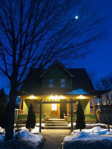 Green house with porch lights glowing at dusk, surrounded by snow and a clear moonlit sky.