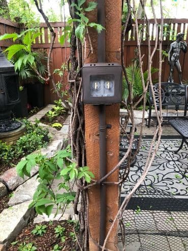 Electrical panel on a wooden post surrounded by lush greenery and outdoor seating area.